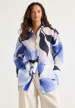 Kvinde med langt krøllet hår iført en løs, knappet skjorte med blå, sorte og hvide abstrakte mønstre og offwhite bukser.