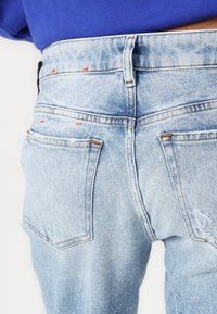 Jeans de mezclilla azul claro con un acabado desgastado y costuras visibles. Presentan dos bolsillos traseros y detalles de costura roja cerca de la cintura.