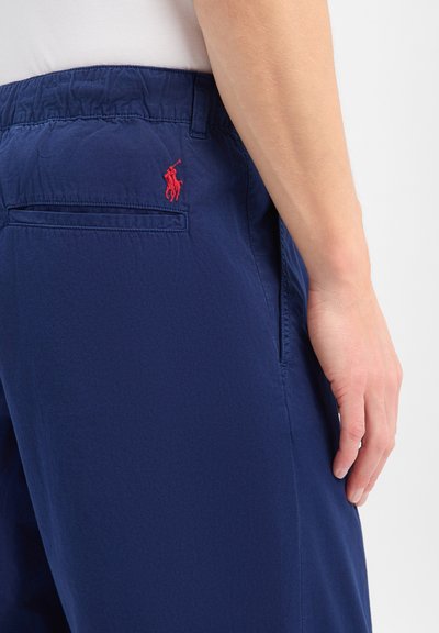 Gros plan d'une personne portant un pantalon décontracté bleu marine avec un logo joueur de polo rouge brodé près de la poche arrière côté droit.