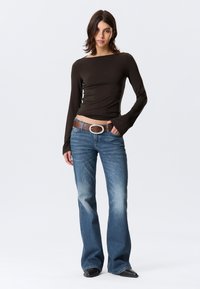 Καφέ μακρυμάνικη μπλούζα με λαιμόκοψη σκάφος· συνδυασμένη με ξεθωριασμένο μπλε τζιν καμπάνα. Καφέ ζώνη με ασημένια στρογγυλή αγκράφα· λεία υφή.