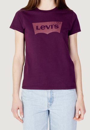 Kobieta w fioletowej koszulce Levi's i jasnoniebieskich jeansach, pokazana od ramion do bioder na jednolitym tle.