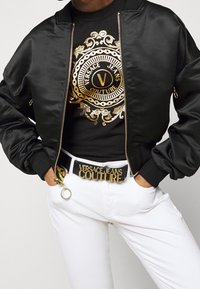 Mustan satiin bomber-takki, jossa on ribbireunuskaulus, yhdistettynä kultakoristeiseen mustaan t-paitaan ja valkoisiin farkkuihin, joissa on rohkea logovyö.