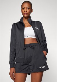 Μαύρο αθλητικό τζάκετ με φερμουάρ, συνδυασμένο με λευκό αθλητικό σουτιέν και μαύρη βερμούδα. Περιέχει το λογότυπο Adidas στο στήθος και στις βερμούδες. Λείο ύφασμα.