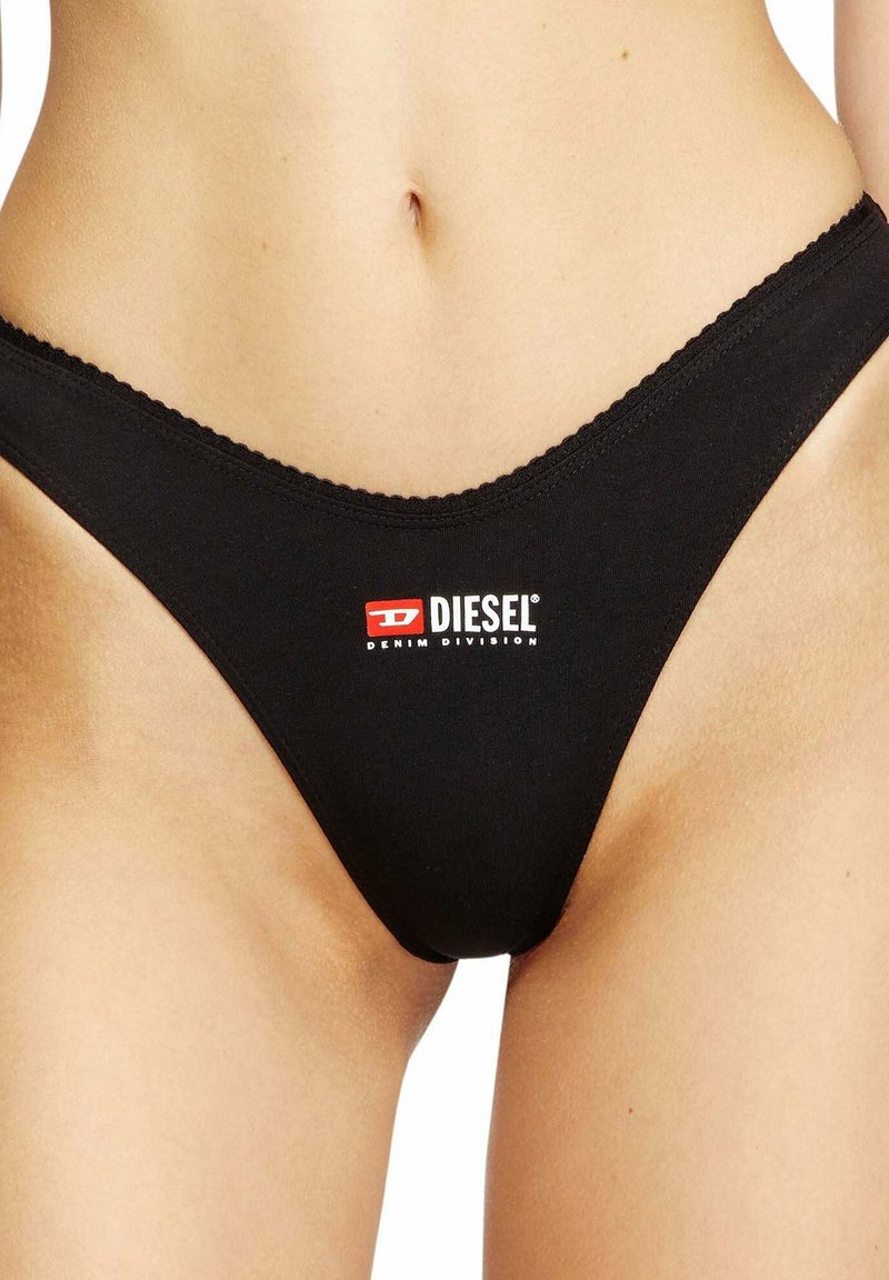 Primer plano de una persona que lleva ropa interior negra Diesel con el logo impreso en el centro delantero, mostrando la parte inferior del torso y la parte superior de los muslos.