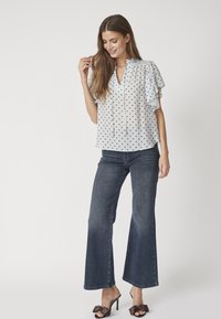Blusa bianca con pois neri, maniche corte a volant e scollatura con lacci, abbinata a jeans a zampa d'elefante blu scuro e sandali neri con tacco.