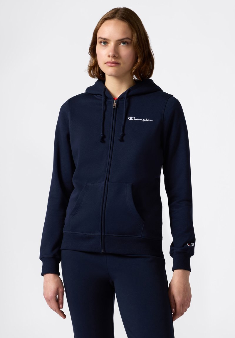 Sudadera con cremallera de color azul marino, hecha de una tela suave y texturizada, con capucha, bolsillos frontales y un logo bordado en el pecho y la manga.