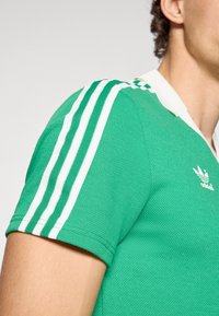 Πράσινο πόλο πουκάμισο με υφή, διακοσμημένο με λευκές τρεις ρίγες στους ώμους και λευκό γιακά. Το λογότυπο της Adidas εμφανίζεται στο στήθος.