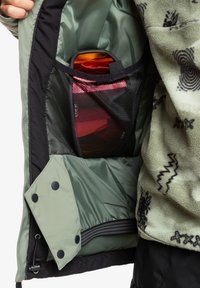 Zelená izolovaná bunda s vnitřním prostorem obsahujícím síťovou kapsu, v níž se nacházejí oranžové a červené brýle, texturovaná fleece tkanina s černými vzory.