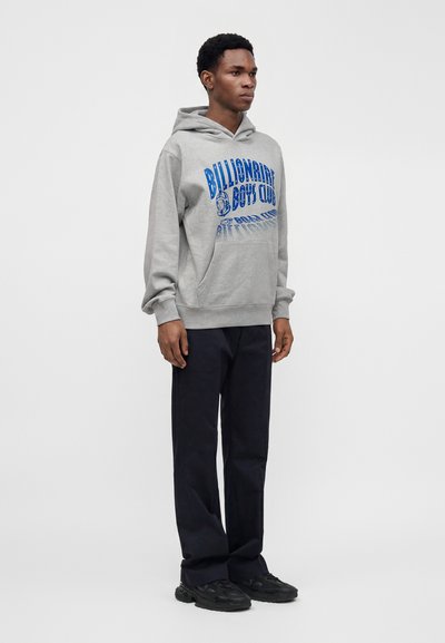 Νέος άνδρας στέκεται φορώντας γκρι φούτερ Billionaire Boys Club, μαύρο παντελόνι και μαύρα παπούτσια σε απλό φωτεινό φόντο.
