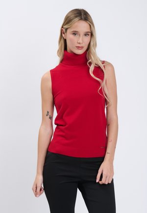 Frau mit langen blonden Haaren trägt einen ärmellosen roten Rollkragenpullover und eine schwarze Hose und steht vor einem schlichten weißen Hintergrund.