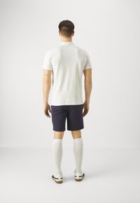 Bílá polo košile, námořnické šortky, bílé ponožky po kolena a bílé boty s hnědými podrážkami. Model obrácen zády k neutrálnímu pozadí.