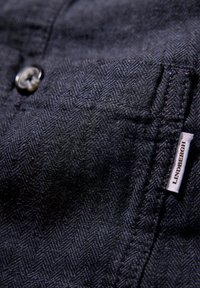 Mörk marinblått tyg med ett fiskbensmönster, en enkel knappstängning och en liten vit etikett med texten "LINDENBERGH." Strukturerad yta.