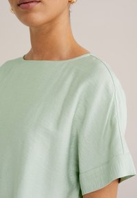 Hele roheline lühikeste varrukatega topp, millel on lai kaelus, sile tekstuur ja peen sära. Omab rullitud varrukaid ja lõdva istuvust.