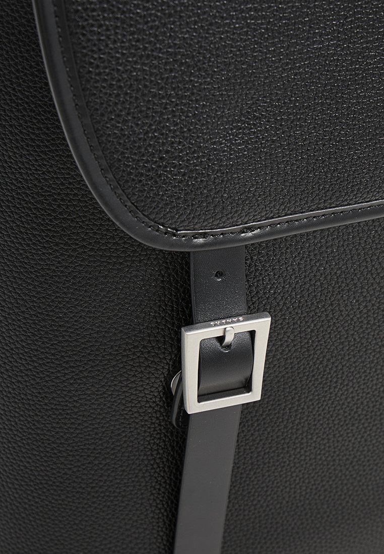 Detailaufnahme einer schwarzen, strukturierten Lederhandtaschenklappe, die mit einem glatten Lederband und einer quadratischen silbernen Metallschnalle gesichert ist.