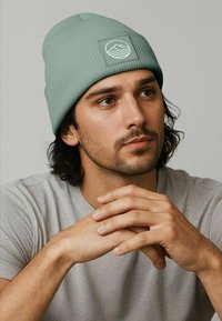 Light green ribbed knit beanie featuring a square patch with mountain and wave graphics. Worn on a model against a neutral background.