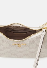 Beżowa torebka materiałowa z subtelnym wzorem monogramu MK, złotym zamkiem błyskawicznym i logo; posiada wewnętrzne kieszonki oraz pasek na nadgarstek.