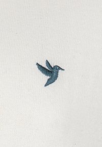 Hvidt stof med en fin strikstruktur, der har et lille teal broderet kolibri-motiv på venstre side.