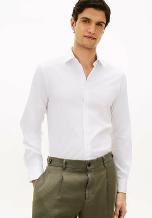 Man wearing a white long-sleeve button-up shirt tucked into olive green pleated trousers with hands in pockets against white background.