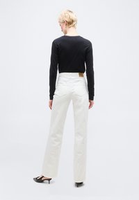 Άτομο με κοντά ξανθά μαλλιά, φορώντας μαύρη μπλούζα με μακριά μανίκια, λευκό τζιν ψηλόμεσο και μαύρα παπούτσια με χαμηλό τακούνι, στέκεται μπροστά σε λευκό φόντο.