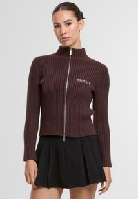 Brązowy prążkowany sweter zapinany na zamek z wysokim kołnierzem, z napisem "ANOTHER" w jasnym kolorze nici. Noszony z czarną plisowaną mini spódniczką.