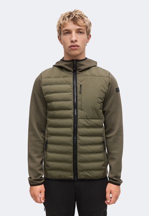 Junger Mann mit kurzen blonden Haaren, der eine grüne gesteppte ärmellose Jacke über einem grünen langärmeligen Hoodie trägt, stehend vor einem schlichten Hintergrund.