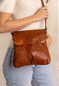 Brun læder crossbody taske med flap-lukning, blød tekstur og subtil kornstruktur. Har en lang justerbar rem og minimal hardware.