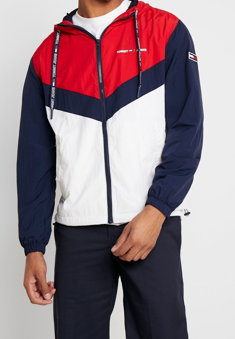 Red, navy, and white windbreaker with a zip front, hood, and elastic cuffs. Features Tommy Jeans logo on the chest and sleeve. Lightweight material.
