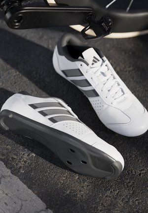 Witte fietsschoenen met zwarte zolen en drie zwarte strepen, neergelegd op asfalt naast een zwart fiets pedaal.