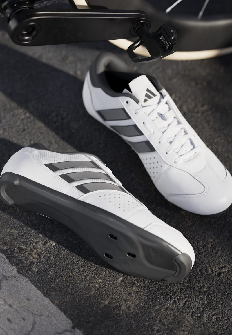 Bílé cyklistické boty s černými podrážkami a třemi černými pruhy, umístěné na asfaltu poblíž černého pedálu bicyklu.