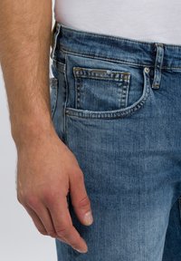 Blauwe denim jeans met een lichte wassing, voorzien van vijf zakken, zichtbare stiksels en een tailleband met riemlussen. Hand rustend op de zak.
