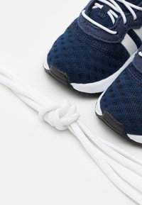 Pari kenkästä tummansiniset tennarit valkoisella nauhalla vieressä erilliset valkoiset kengännauhat solmittuna solmuun valkoisella pinnalla.