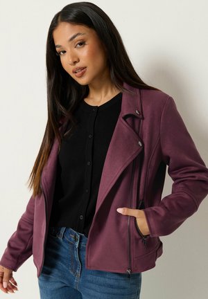 Mujer con cabello largo y oscuro que lleva una chaqueta morada con cremallera sobre una blusa negra con botones y vaqueros azules, de pie contra un fondo claro y sencillo.
