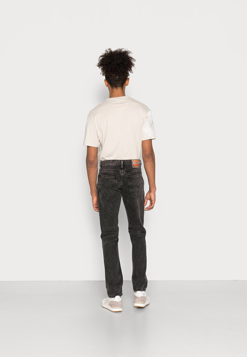 Beige T-Shirt mit bedrucktem Ärmel, kombiniert mit schwarzen Jeans. Das Outfit hat eine lässige Passform und wird durch weiße Sneakers ergänzt.