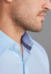 Himmelblaues, strukturiertes Hemd mit einem geknöpften Kragen, der mit einem schwarzen Muster gefüttert ist. Nahaufnahme des Kragendetails.
