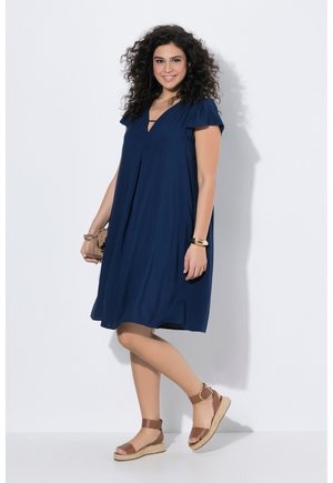 Kvinde med krøllet hår iført en mørkeblå knælang kjole, brune sandaletter med ankelrem og med en lille beige taske i hånden.