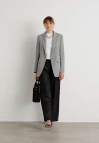 Světle šedý blazer, bílý top a široké černé kalhoty. Černá kabelka s pevnou strukturou. Špičaté béžové podpatky. Jednoduché stříbrné šperky.