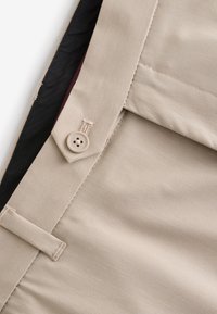 Tessuto beige con un bottone cucito e dettaglio ad asola, mostrando parte del bordo piegato di un indumento e la fodera interna nera.