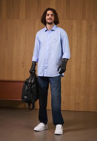 Lyseblå langermet skjorte, svart lærveske, mørke jeans, hvite joggesko; funksjoner inkluderer knappdetaljer og svarte hansker.