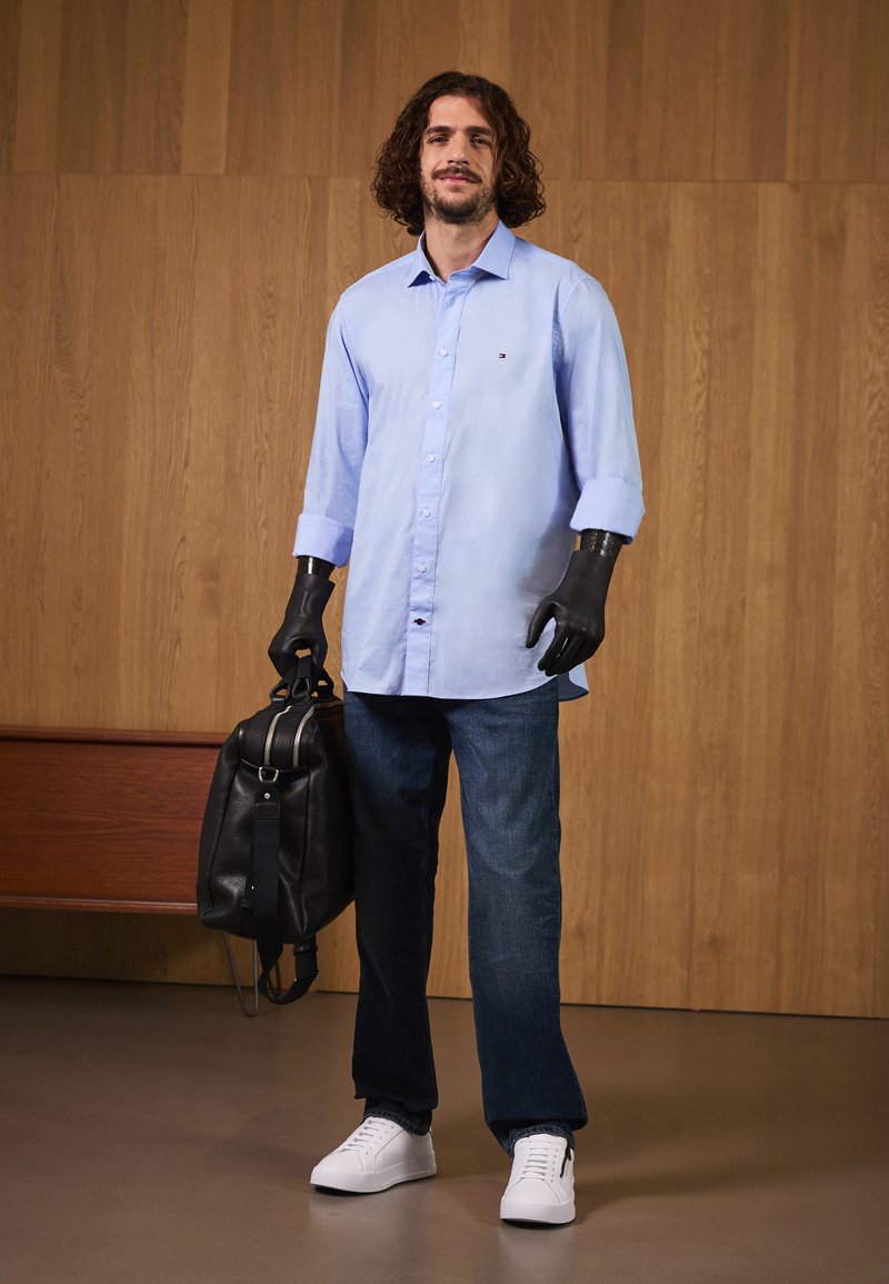 Lyseblå langermet skjorte, svart lærveske, mørke jeans, hvite joggesko; funksjoner inkluderer knappdetaljer og svarte hansker.