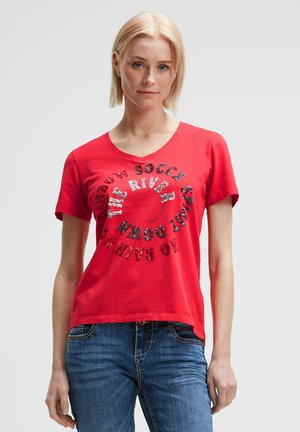 Blonde woman wearing a red short-sleeve T-shirt with circular black and white text and blue jeans, standing against a plain light background.