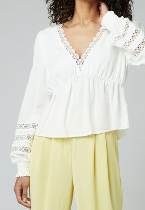 Blouse blanche avec un col en V, des bordures en dentelle et des manches longues bouffantes avec des accents en dentelle transparente. Associée à un pantalon plissé jaune clair.