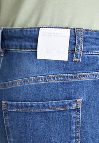 Blaue Jeans aus Denim mit einem weißen Stoffetikett, auf dem "MARC CAIN" und Pflegehinweise stehen, akzentuiert mit orangen Nähten und einer Gesäßtasche.