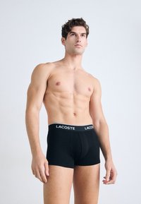 Spiergevende man in zwarte Lacoste boxershorts, staand tegen een effen lichte achtergrond, met een lichte blik omhoog en opzij.