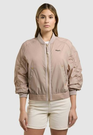 Naine, kes kannab beeži bomber-tüüpi jopet voltse käistega ja heledaid lühikesi pükse, seisab ühevärvilise heleda tausta ees.