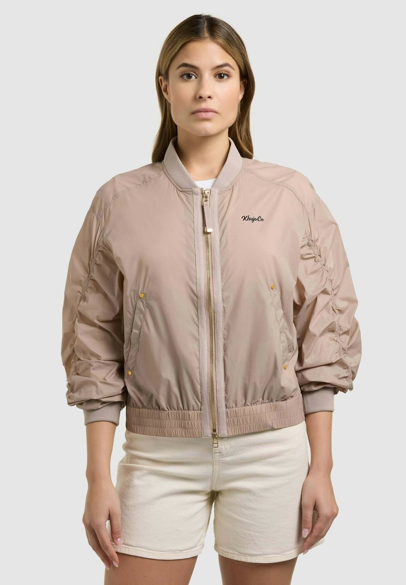 Femme portant une veste bomber beige à manches froncées et un short de couleur claire, debout devant un fond uni clair.