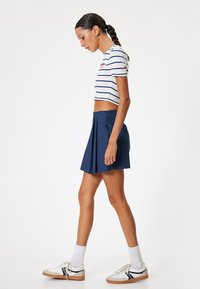 Weißes, kurzes gestreiftes Oberteil mit blauen Linien, kombiniert mit einem marineblauen, plissierten Rock, weißen Socken und off-white Sneakers mit schwarzen Akzenten.