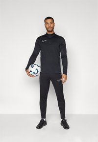 Schwarzes Nike Sportset, bestehend aus einem Reißverschluss-Oberteil und enganliegenden Hosen. Strukturierter Stoff, dezente Logoakzente und ein Fußball, der in einer Hand gehalten wird.