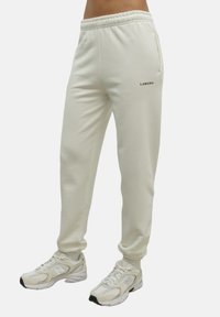 Jasne, kremowe spodnie dresowe o luźnym kroju, z elastycznym paskiem w talii, kieszeniami bocznymi oraz czarnym logo "LAMANIA". Miękka tekstura.