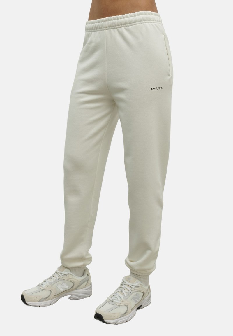 Jasne, kremowe spodnie dresowe o luźnym kroju, z elastycznym paskiem w talii, kieszeniami bocznymi oraz czarnym logo "LAMANIA". Miękka tekstura.