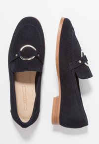 Mocassini in suede blu navy con punta arrotondata, decorazioni in metallo argentato e un tacco basso marrone. Texture liscia con dettagli cuciti.